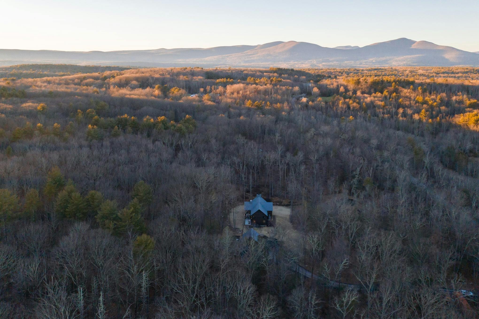 beautiful drone shot of a large darkly painted house nestled in woods with the Catskills in the background